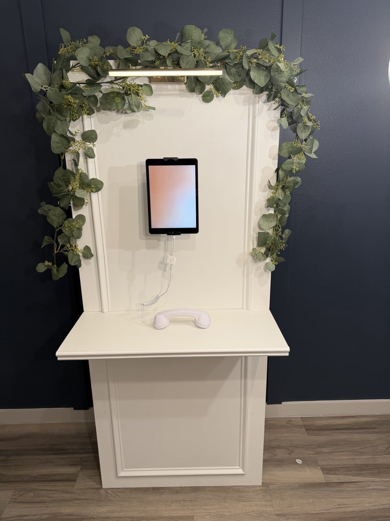 The Memory Wall in daylight — cream paneled wall, brass picture light, iPad guestbook screen and white handset, framed by eucalyptus garland