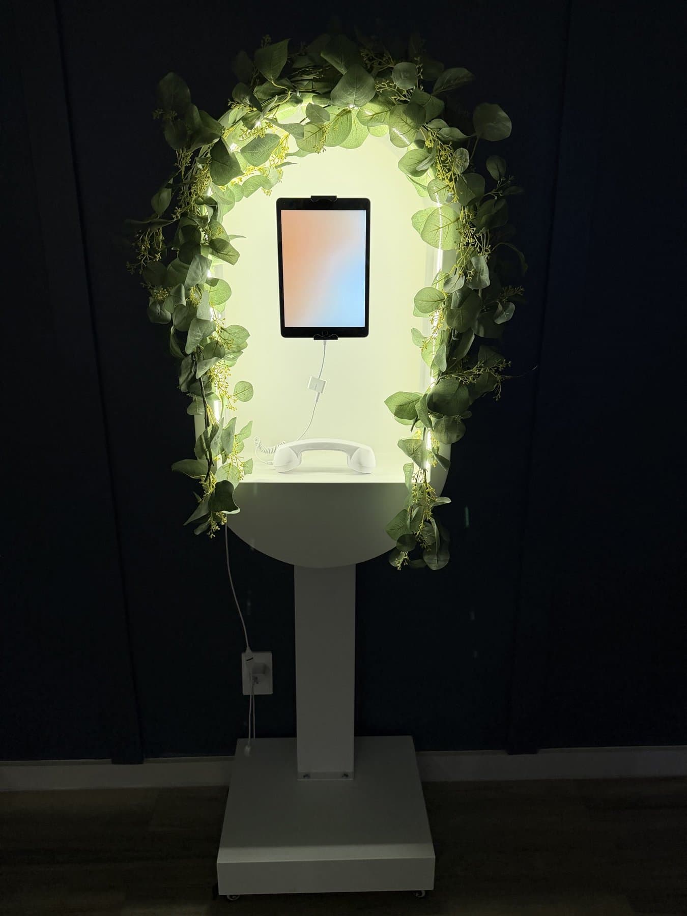 The Video Guestbook kiosk at night — a cream arched stand glowing from its backlit frame, with an iPad recording screen and white handset wrapped in eucalyptus and fairy lights