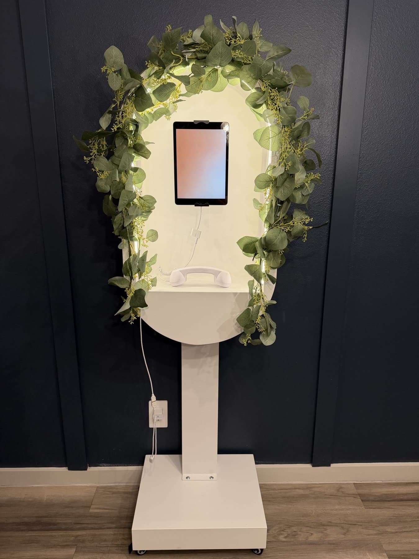 The Video Guestbook kiosk straight on — a cream arched stand with a backlit frame, iPad recording screen and white handset, wrapped in eucalyptus garland against a navy wall